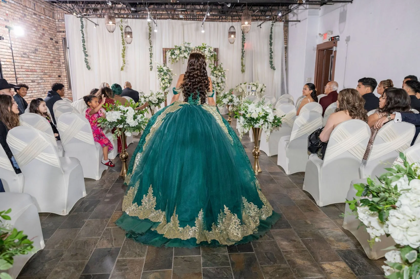 A young woman with long curly hair wearing an ornate green and gold ball gown stands in the center of a decorated room with guests seated on either side, facing her, during a celebration or event.