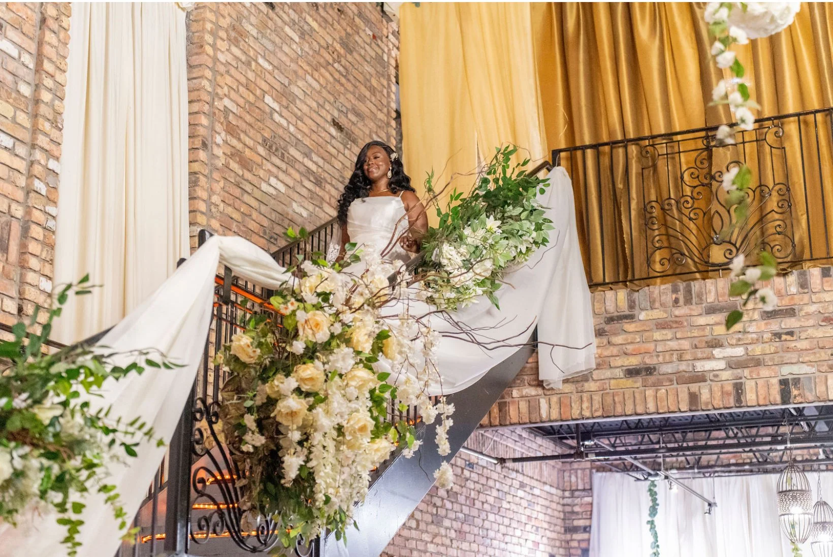 Bride in a white wedding gown standing on a staircase decorated with white and cream flowers and green foliage, inside a venue with exposed brick walls, gold curtains, and decorative wrought iron railing.