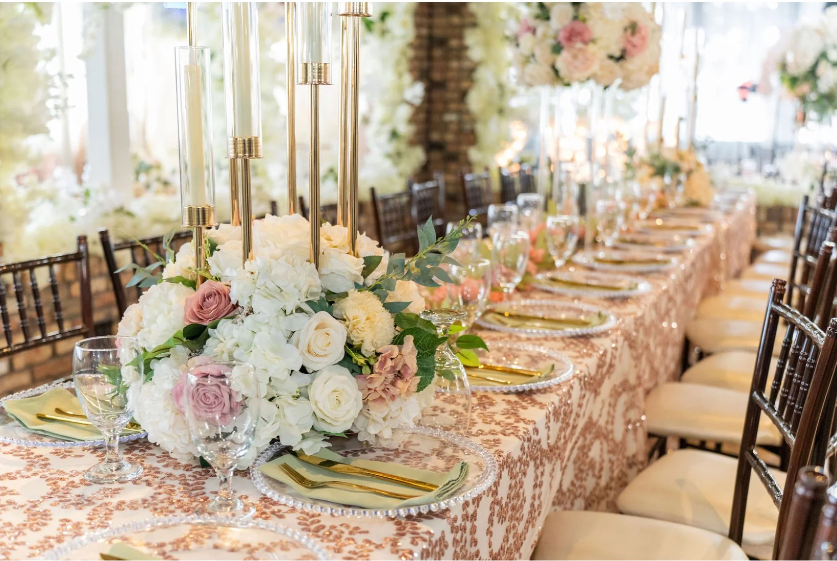 A long wedding reception table decorated with floral centerpieces and gold candle holders, set with glassware, gold utensils, pink napkins, and ornate plates, in a light-filled venue with a brick wall background.