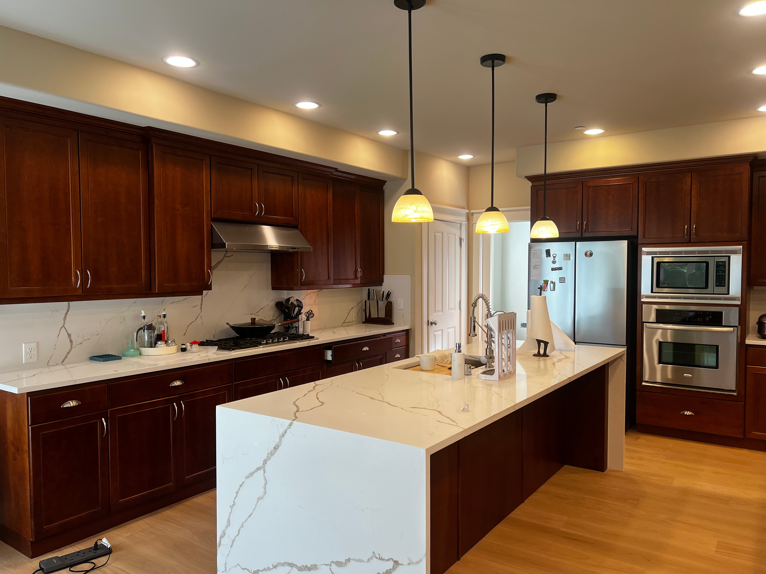 Modern kitchen with dark wood cabinets, white marble countertops, and a kitchen island. The kitchen has stainless steel appliances, including a refrigerator, microwave, and oven. Three pendant lights hang over the island. The floor is wood, and the c