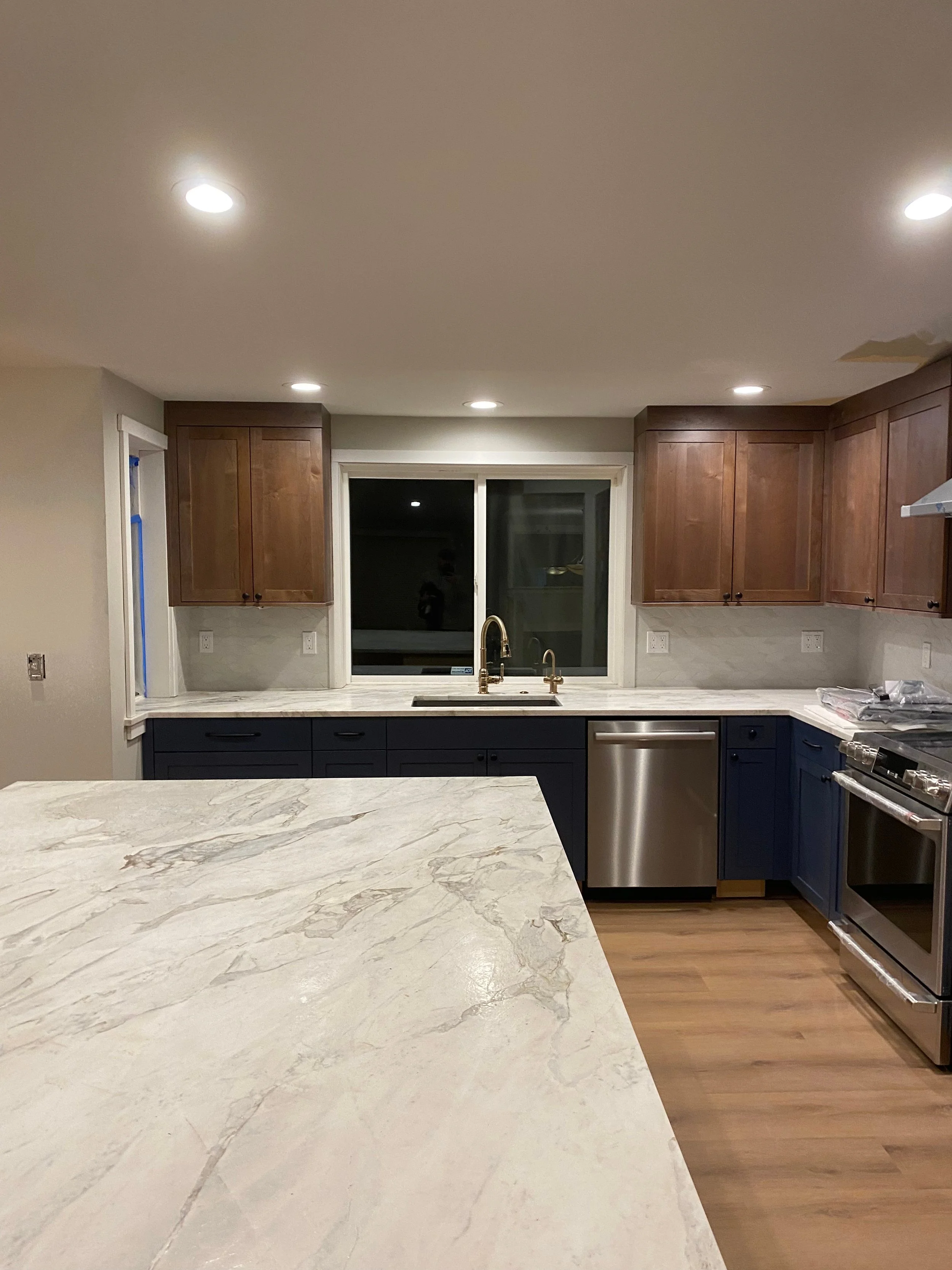 Modern kitchen with marble countertops, wooden upper cabinets, dark blue lower cabinets, stainless steel dishwasher and oven, and a large window above the sink.