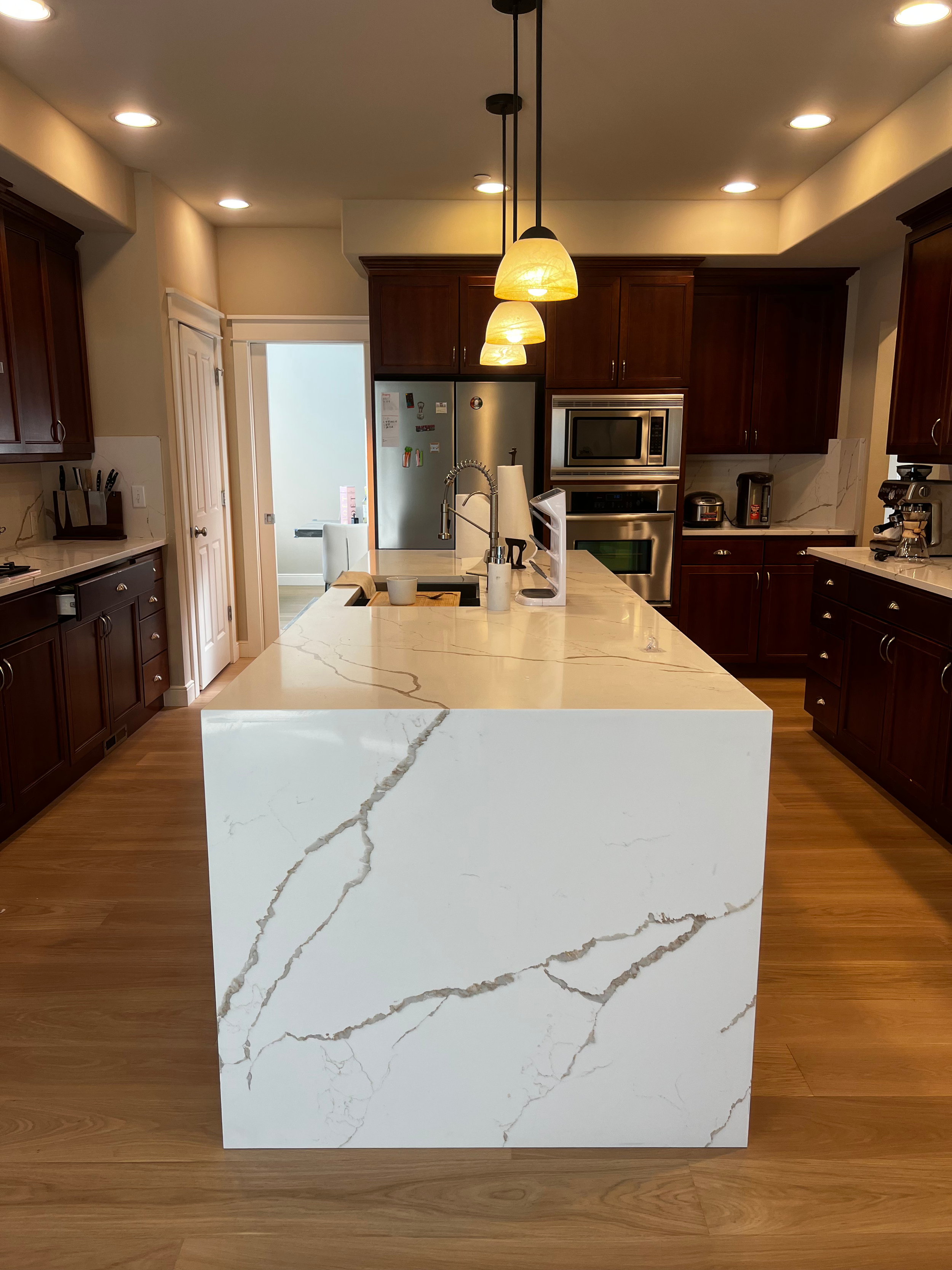 Modern kitchen with a large white marble island with gray veining, dark wood cabinets, stainless steel appliances, and pendant lights.