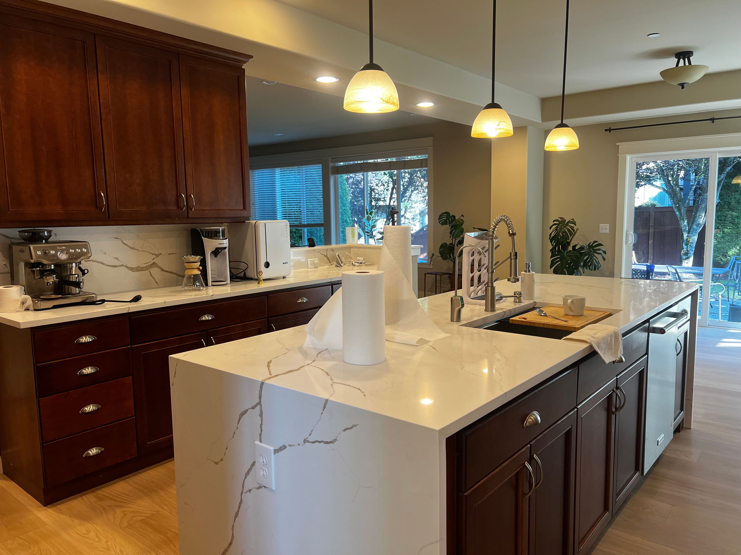 Modern kitchen with marble island, dark wood cabinets, pendant lights, and sliding glass door leading to a backyard with trees.