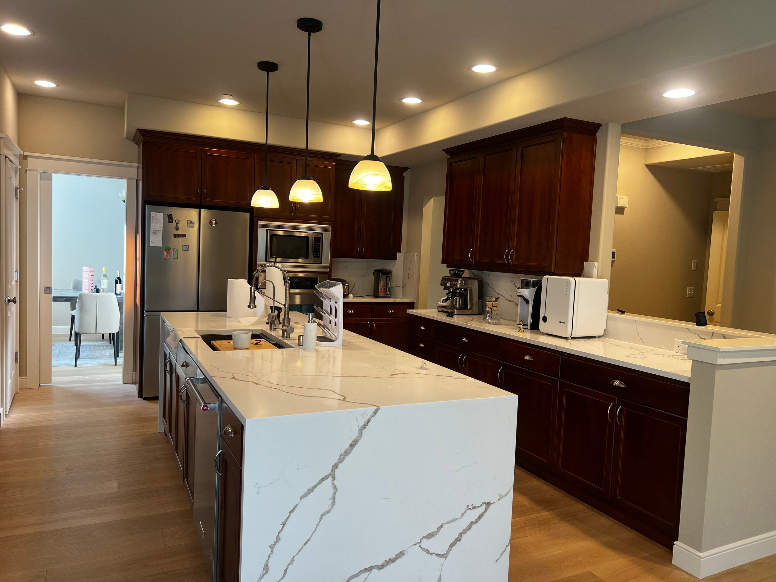 Modern kitchen with dark wood cabinets, white marble countertops, and stainless steel appliances. Pendant lights hang above the island, and a doorway leads to a dining area with a white table and chairs.
