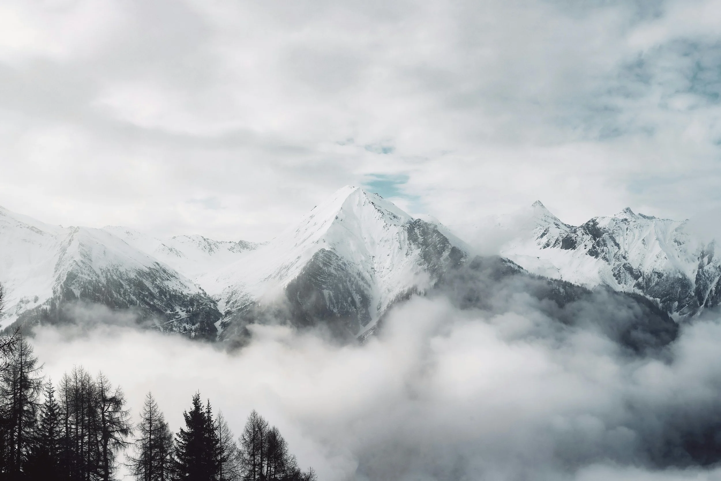 Snow-capped mountain range under cloudy sky, with grassy hills in the foreground.