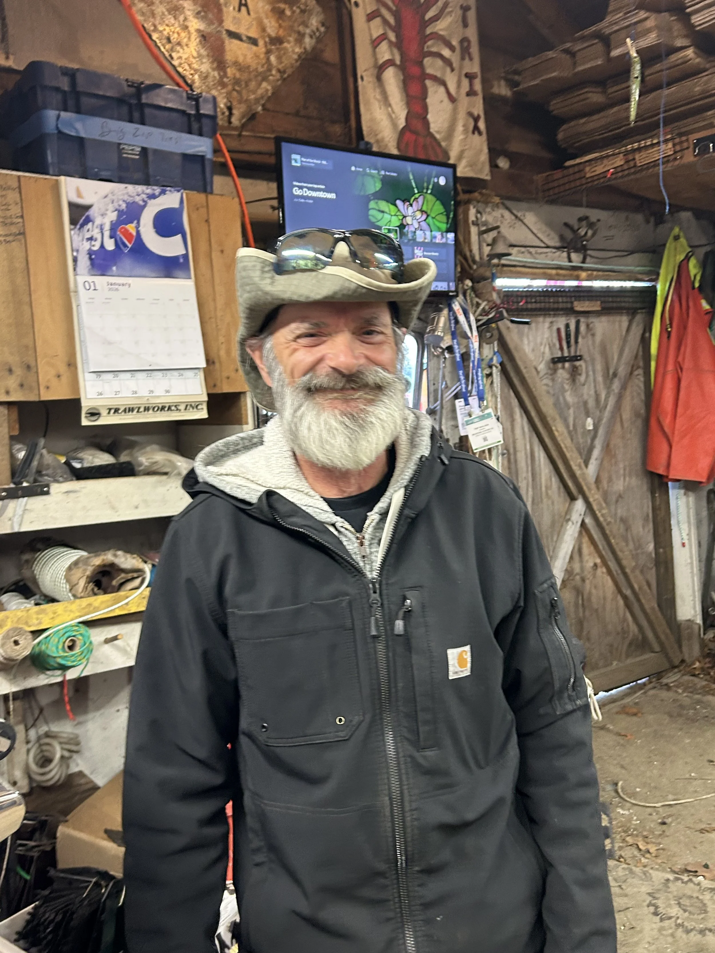 A smiling man with gray hair, a gray beard, wearing a cowboy hat, sunglasses, a black Carhartt jacket, and a gray hoodie inside a workshop or shed.