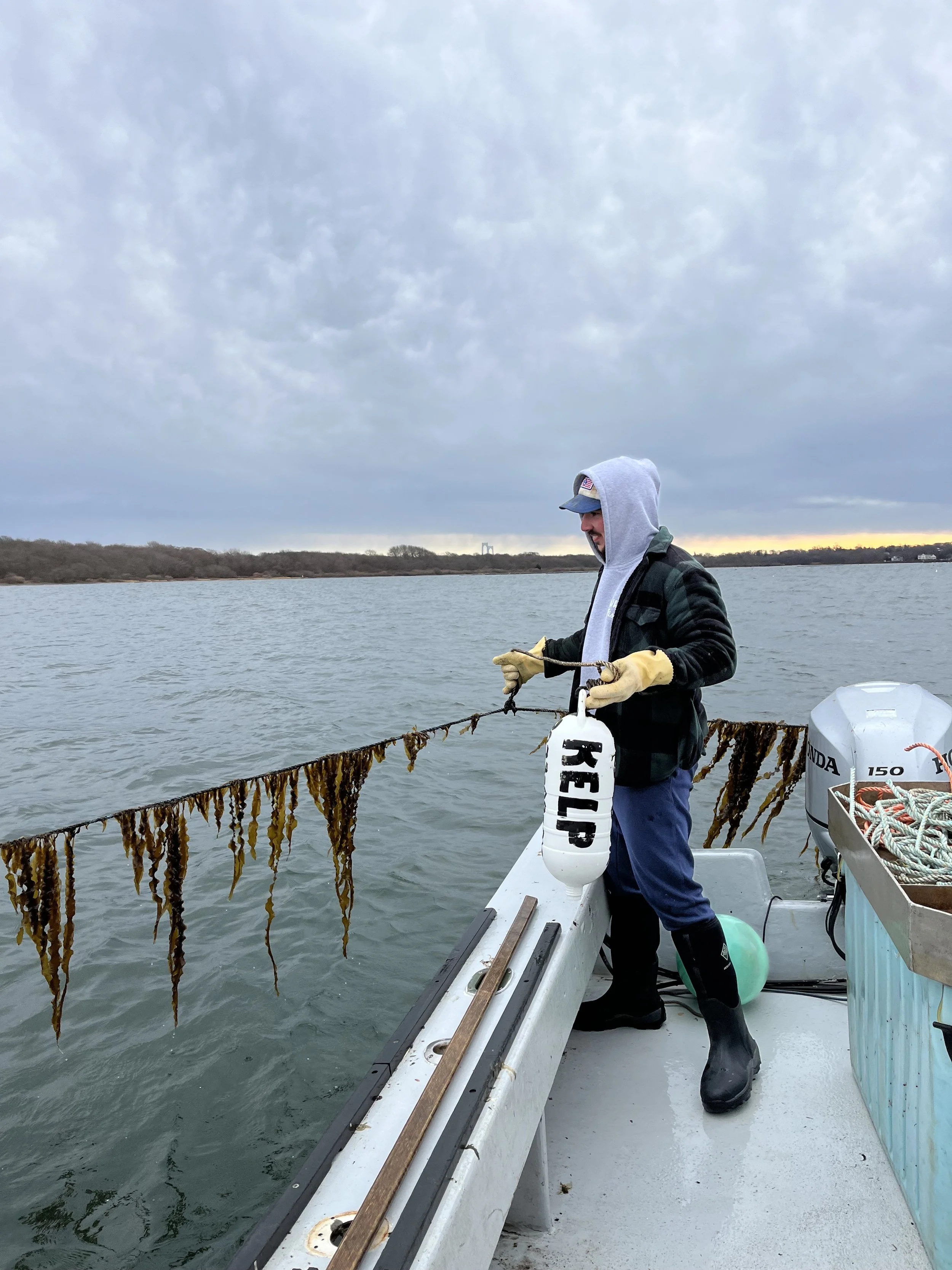 Man on a boat fishing in a body of water during cloudy weather, wearing a hoodie, jacket, gloves, and boots, with seaweed hanging from the fishing line.