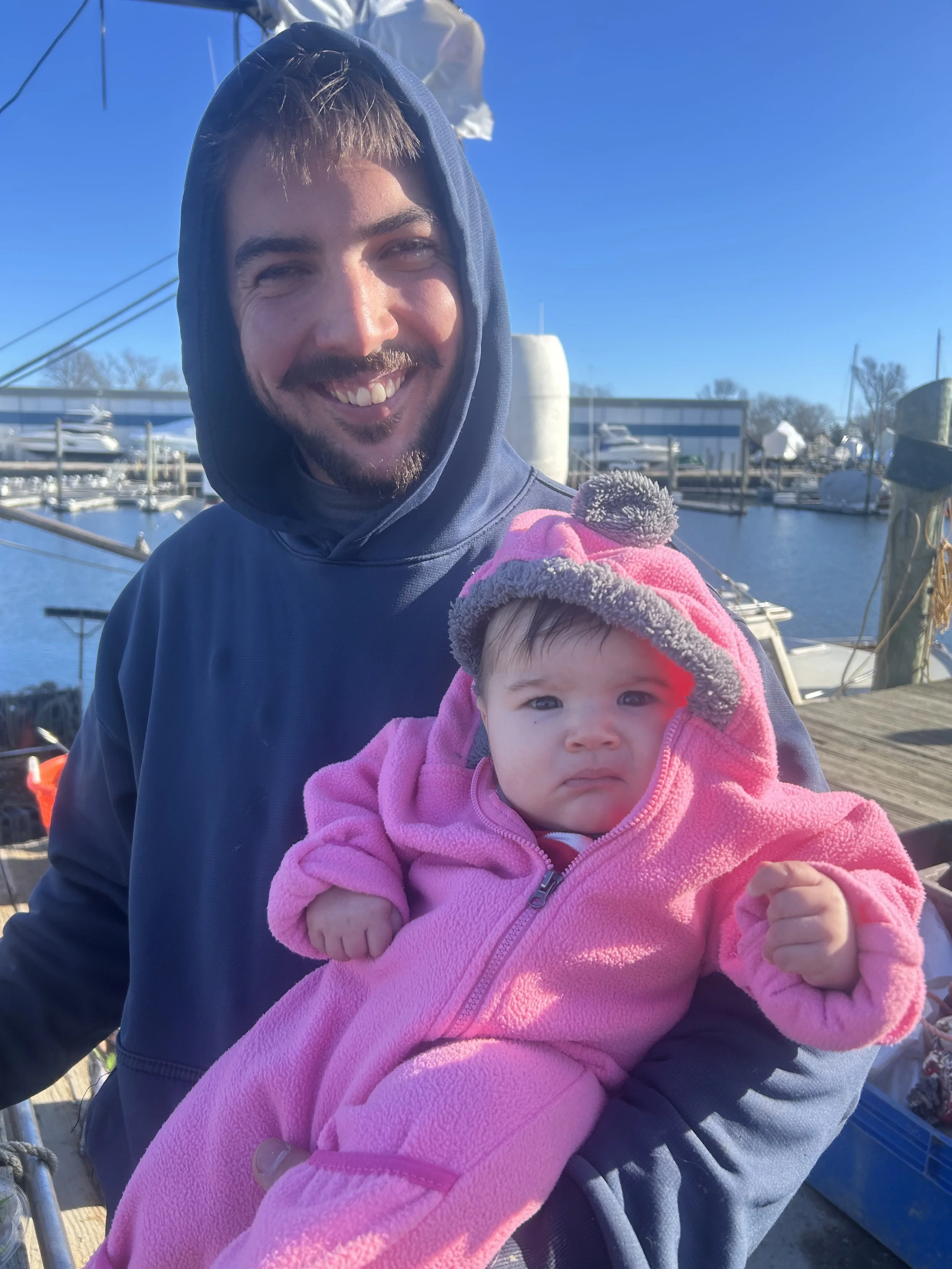 A man holding a baby girl outdoors near a marina with boats, wearing hooded jackets on a sunny day.