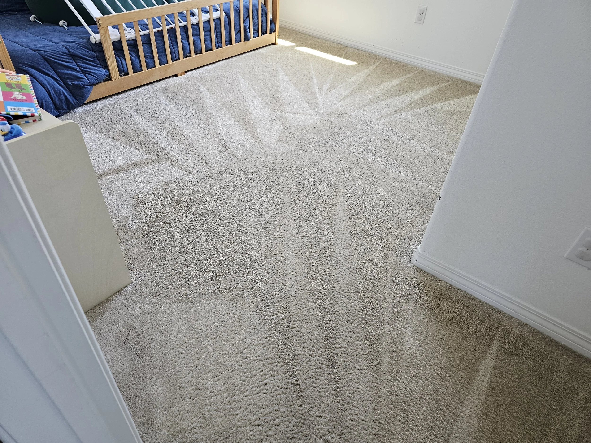 A beige carpet with a sunburst shadow pattern near a white wall and electrical outlet. Part of a wooden bed frame with a blue mattress is visible in the upper left corner, along with some colorful toys or books on a nearby surface.