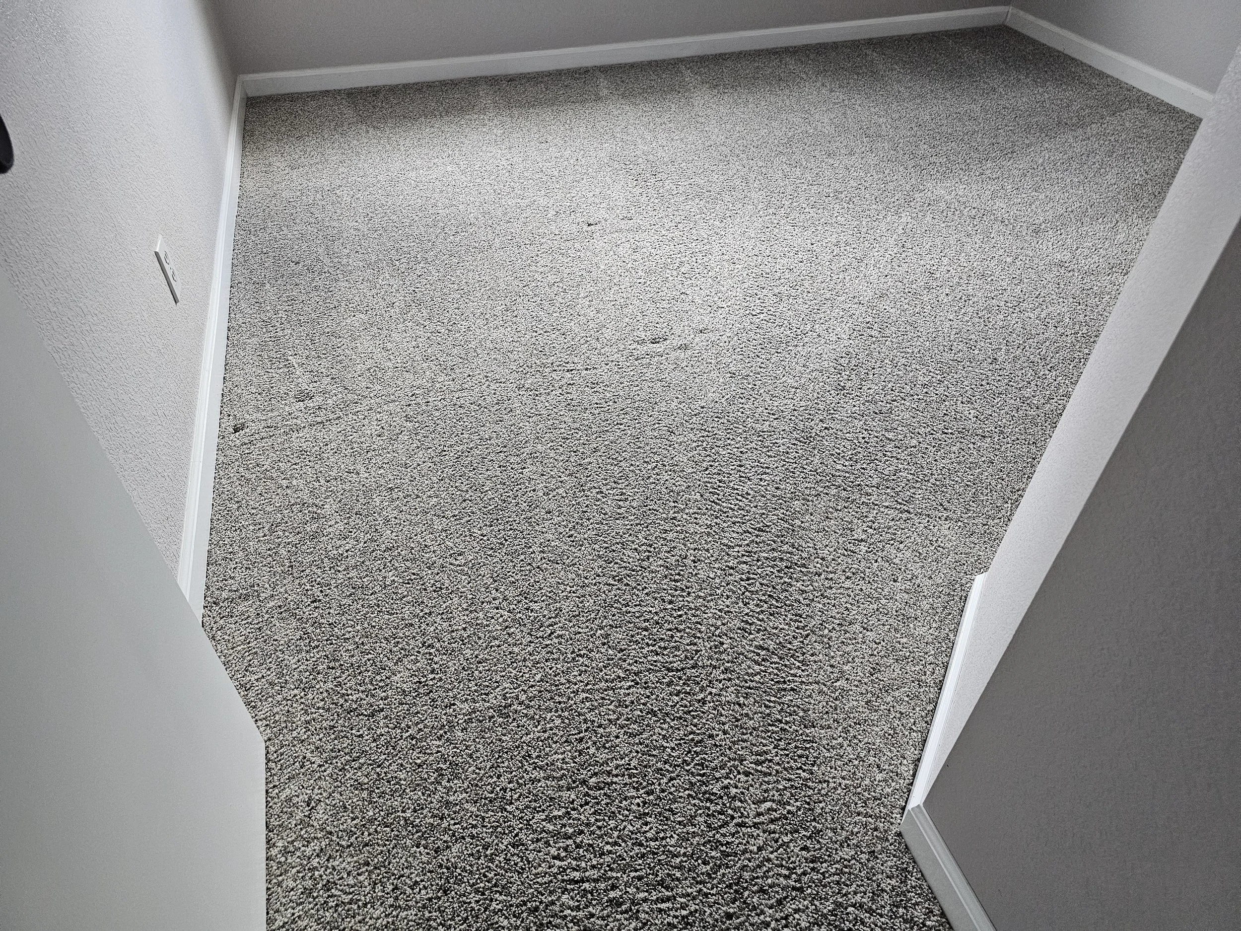 Photo of a small section of a room corner with gray carpet, white baseboards, and white textured walls.