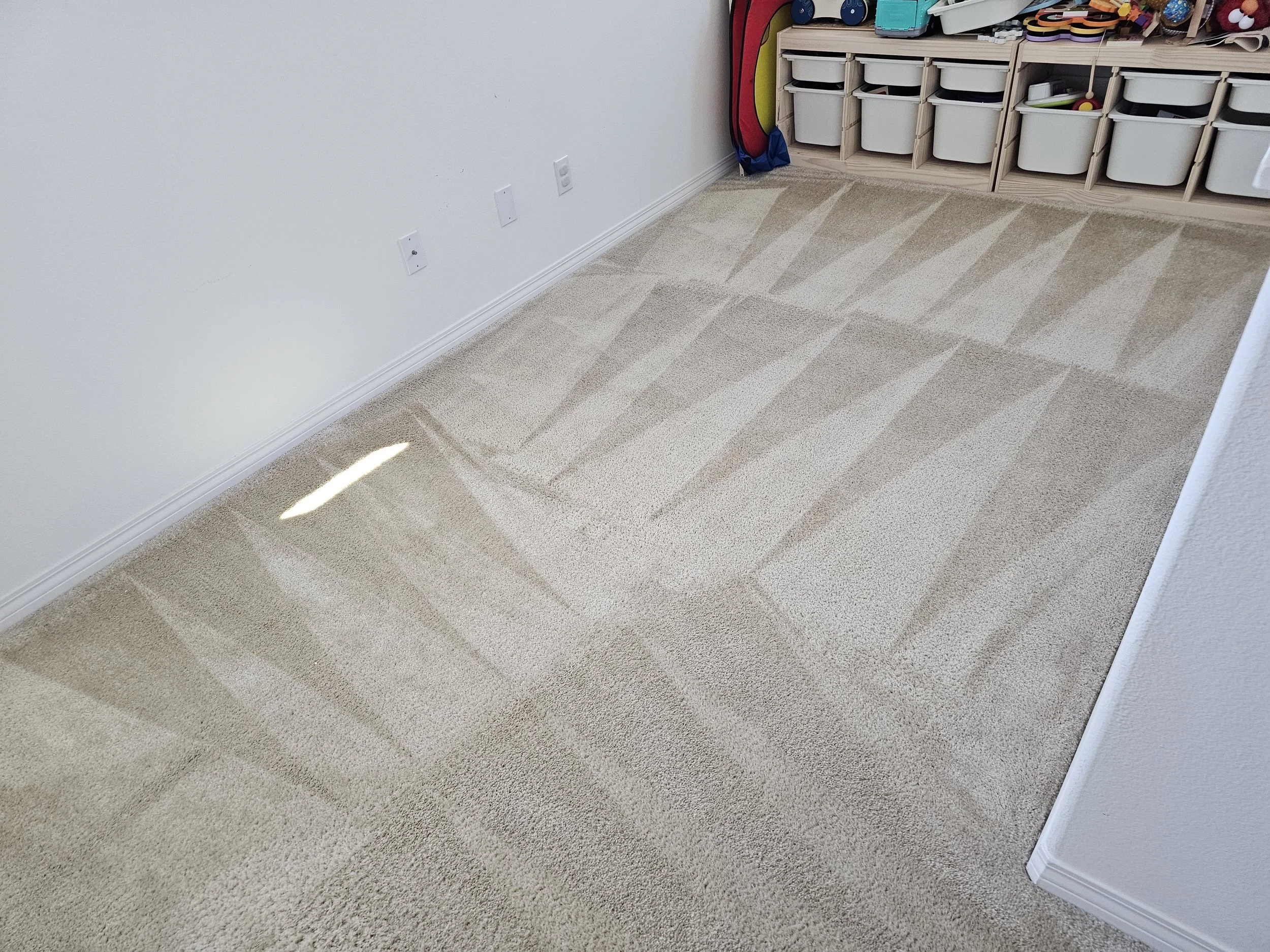 A beige carpeted floor adjacent to a white wall with electrical outlets. To the right, a wooden storage unit filled with plastic bins and some toys, and an assortment of colorful objects on top.
