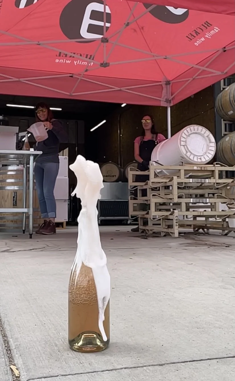 A bottle of beer with foam overflowing from the top, placed on a sidewalk outside a brewery. In the background, two women are preparing beverages under a red umbrella that has the brewery's branding.