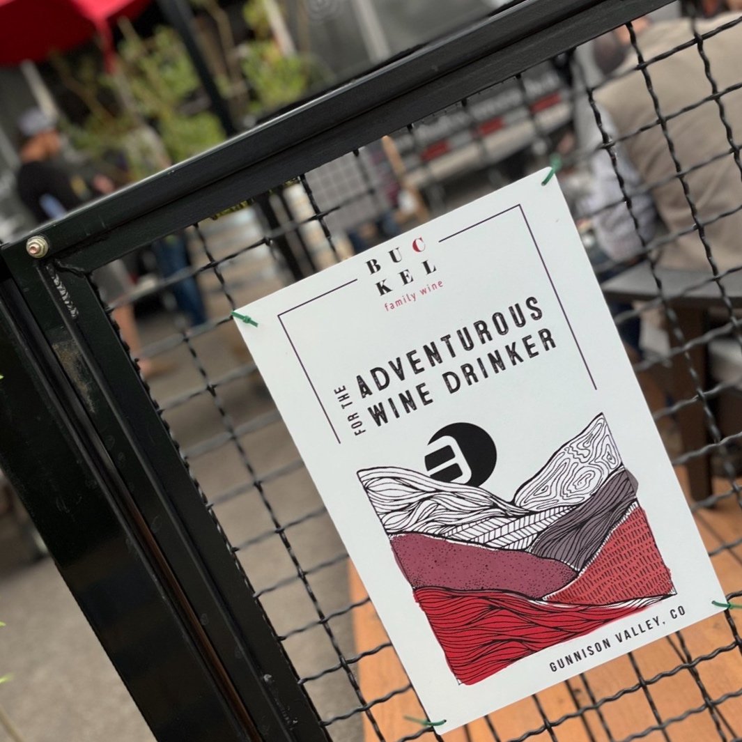 A flyer for the 'Adventurous Wine Drinker' event at Buccle Family Wine in Gunnison Valley, Colorado, attached to a metal fence, with people and outdoor seating in the background.