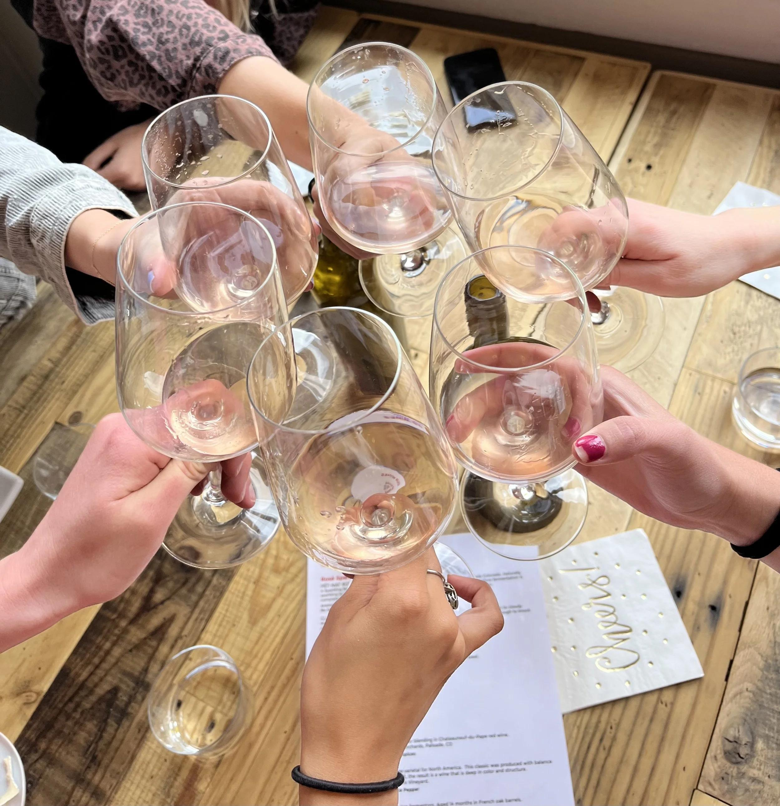 Six people toasting with empty and slightly filled wine glasses over a wooden table.