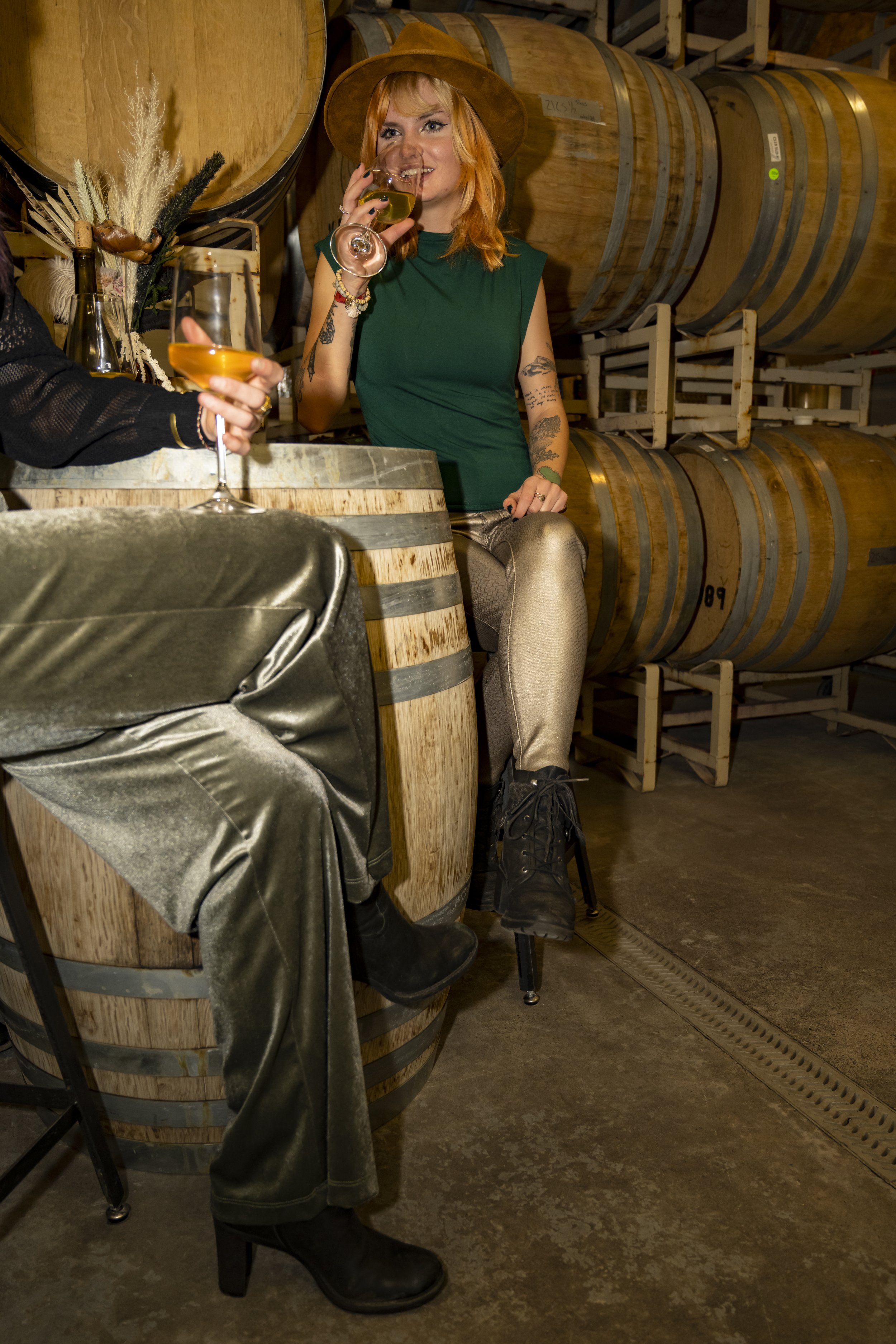 A woman with tattoos and red hair wearing a green sleeveless top, metallic pants, black boots, and a wide-brimmed brown hat, sitting on a barrel in a winery, holding a glass of white wine, with large wine barrels in the background.