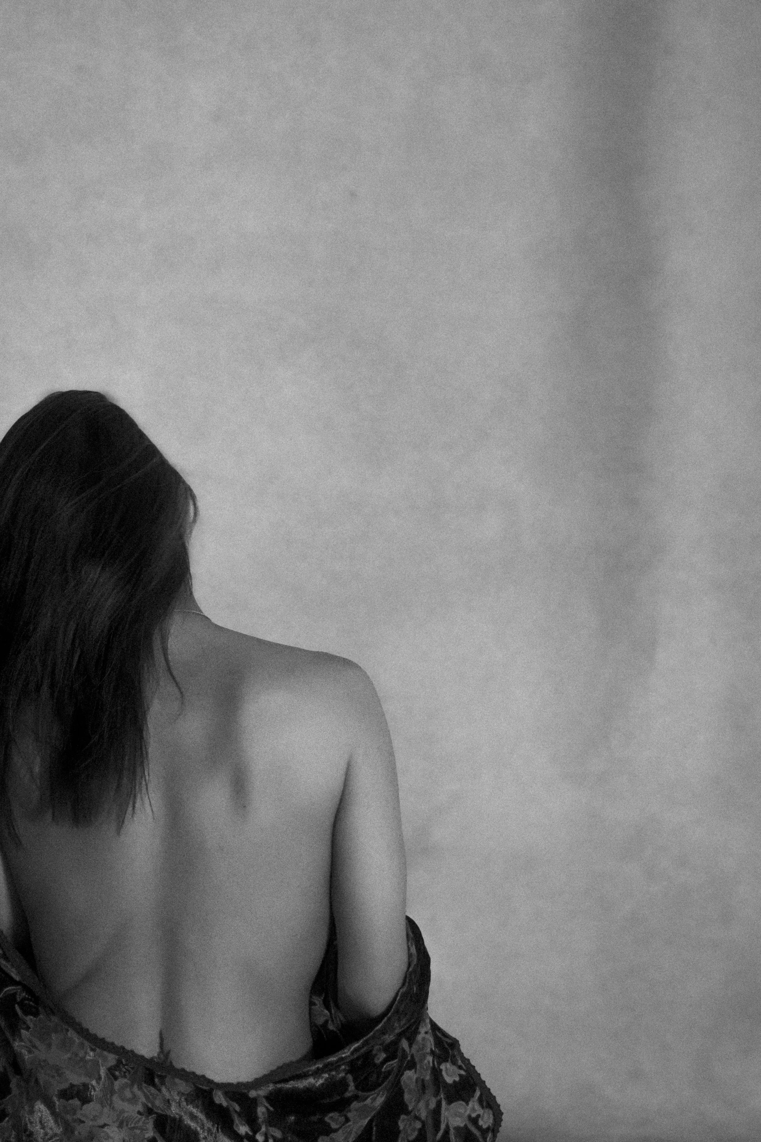 A woman with long hair draped over her shoulder sitting with her back to the camera, wrapped in a patterned cloth in front of a textured light gray background.