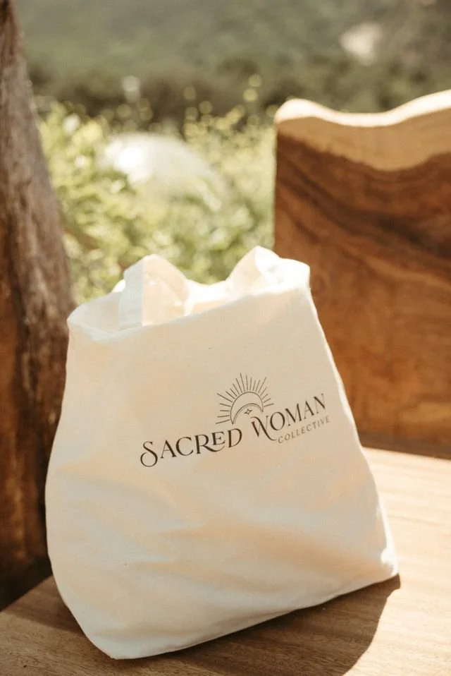 Sacred Woman Collective tote bag resting on a wooden chair outdoors, featuring minimalist Sacred Woman Collective logo design with sun rays.