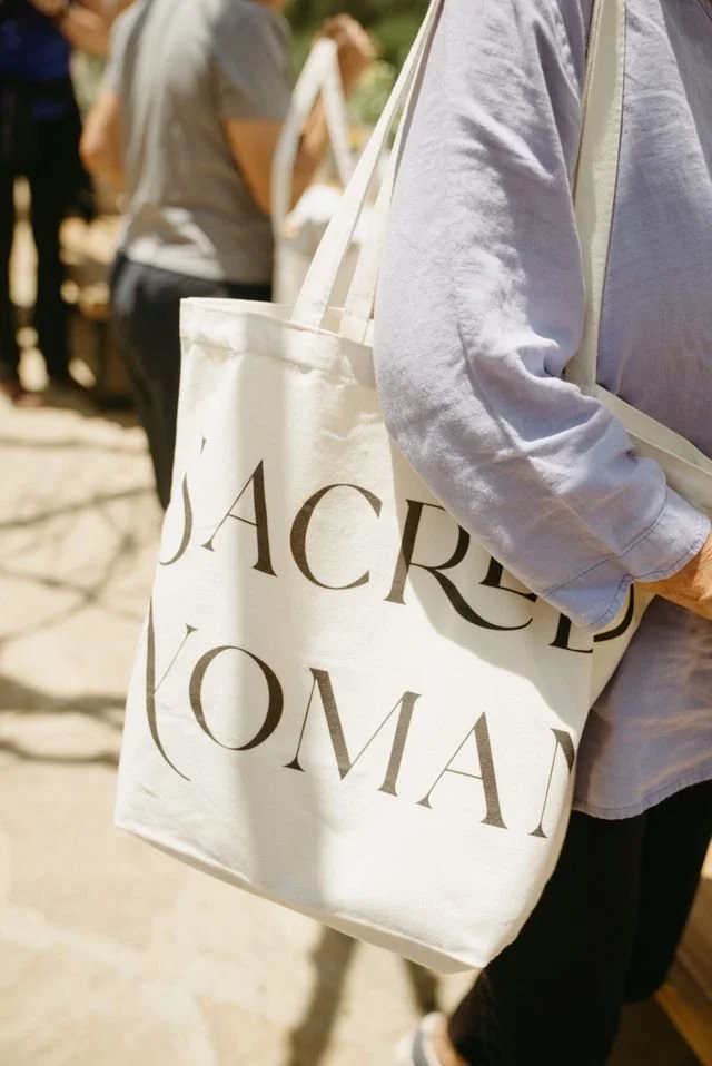 Woman carrying a Sacred Woman Collective tote bag with large bold text design, photographed in natural sunlight
