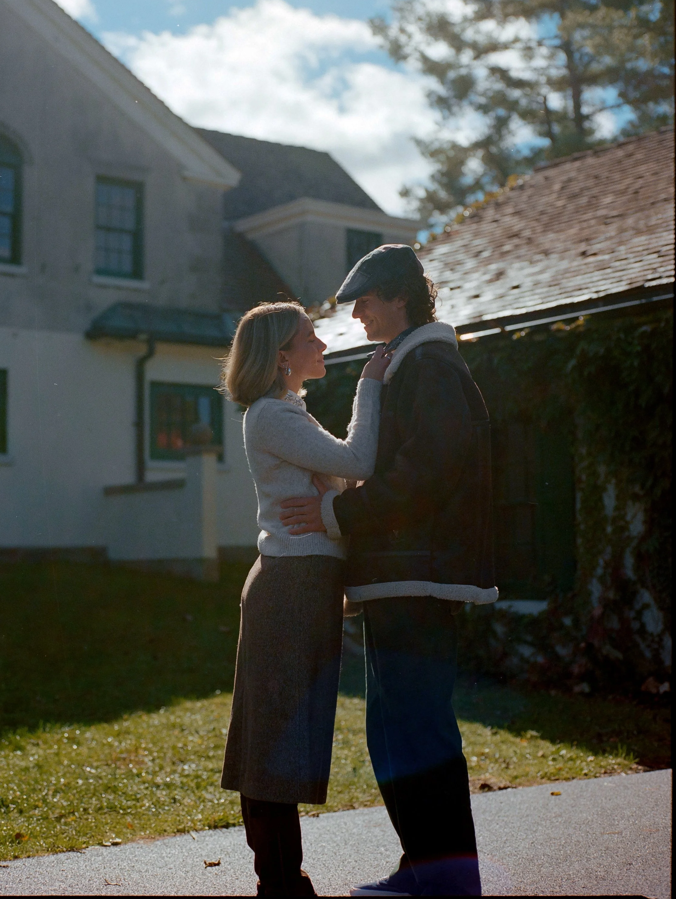A couple standing close to each other outdoors on a sunny day, holding each other's faces and smiling, with houses and trees in the background.