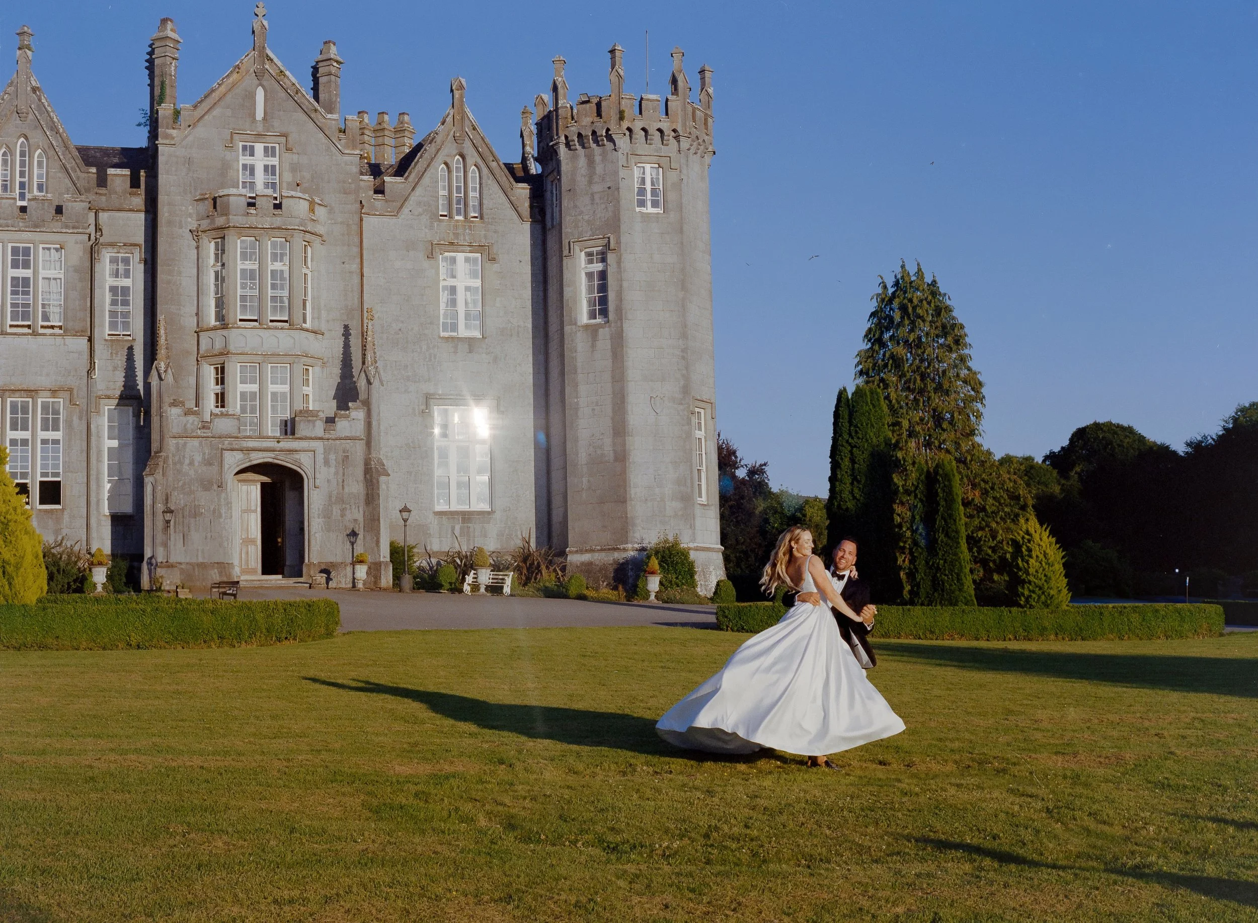A bride and groom dancing on a lawn in front of a large castle-like mansion on a sunny day.