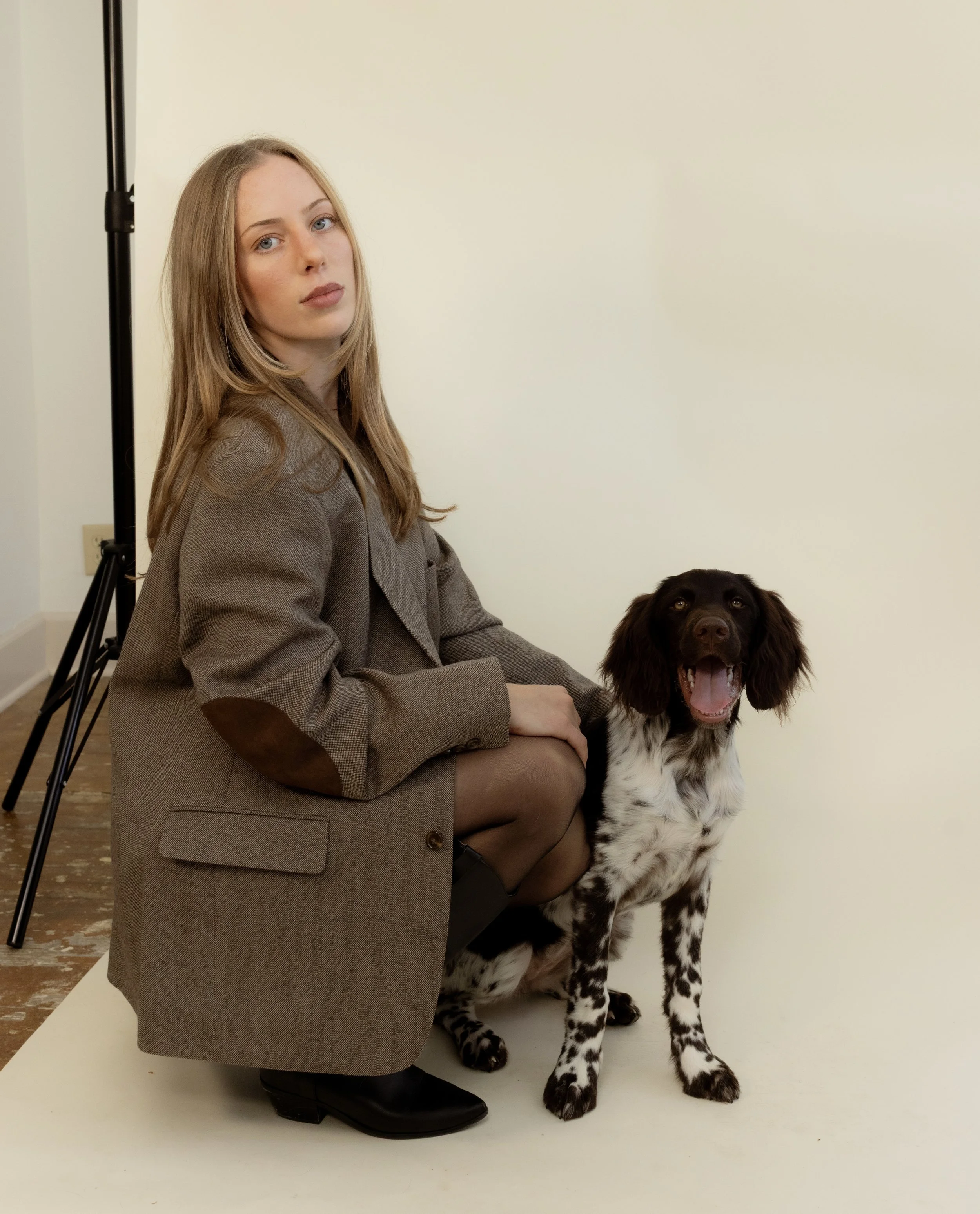 A young woman with long blonde hair wearing a brown coat and black boots, sitting on a white backdrop next to a black and white dog with long ears and a happy open mouth.