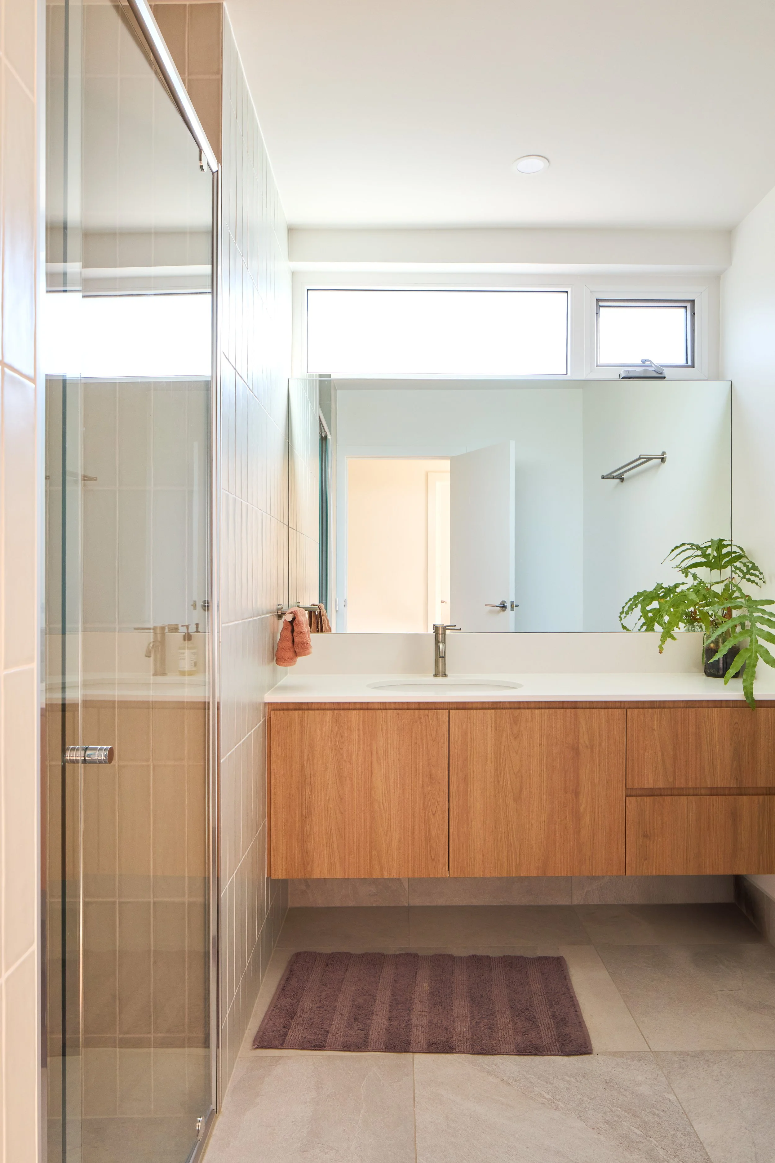 Modern backlit bathroom with wooden vanity, large mirror, potted plant, towel rack, and walk-in shower with glass door.