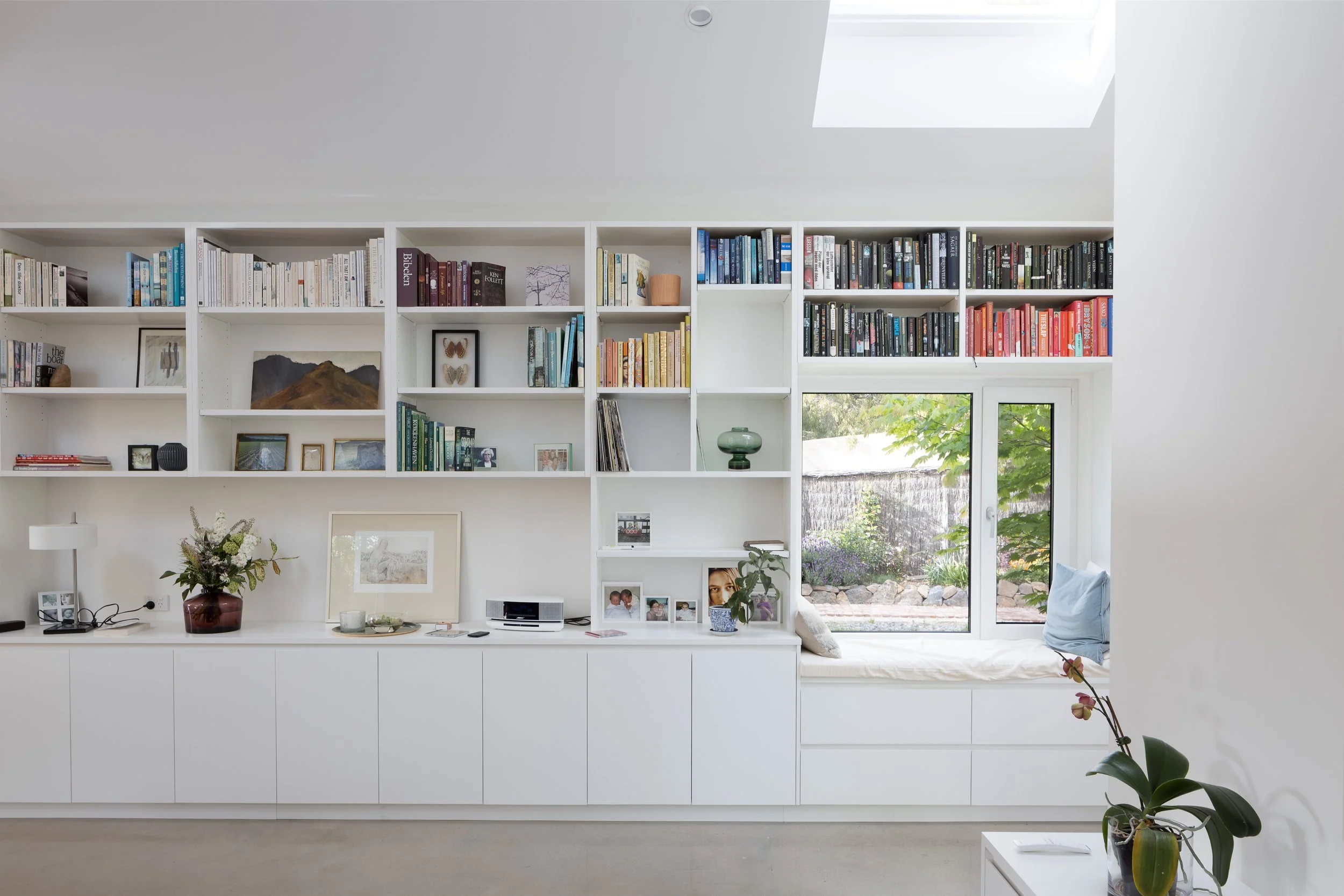 A white built-in bookshelf with various books, photos, and decorative items in a modern living space, with a window seat and large window showing greenery outside.