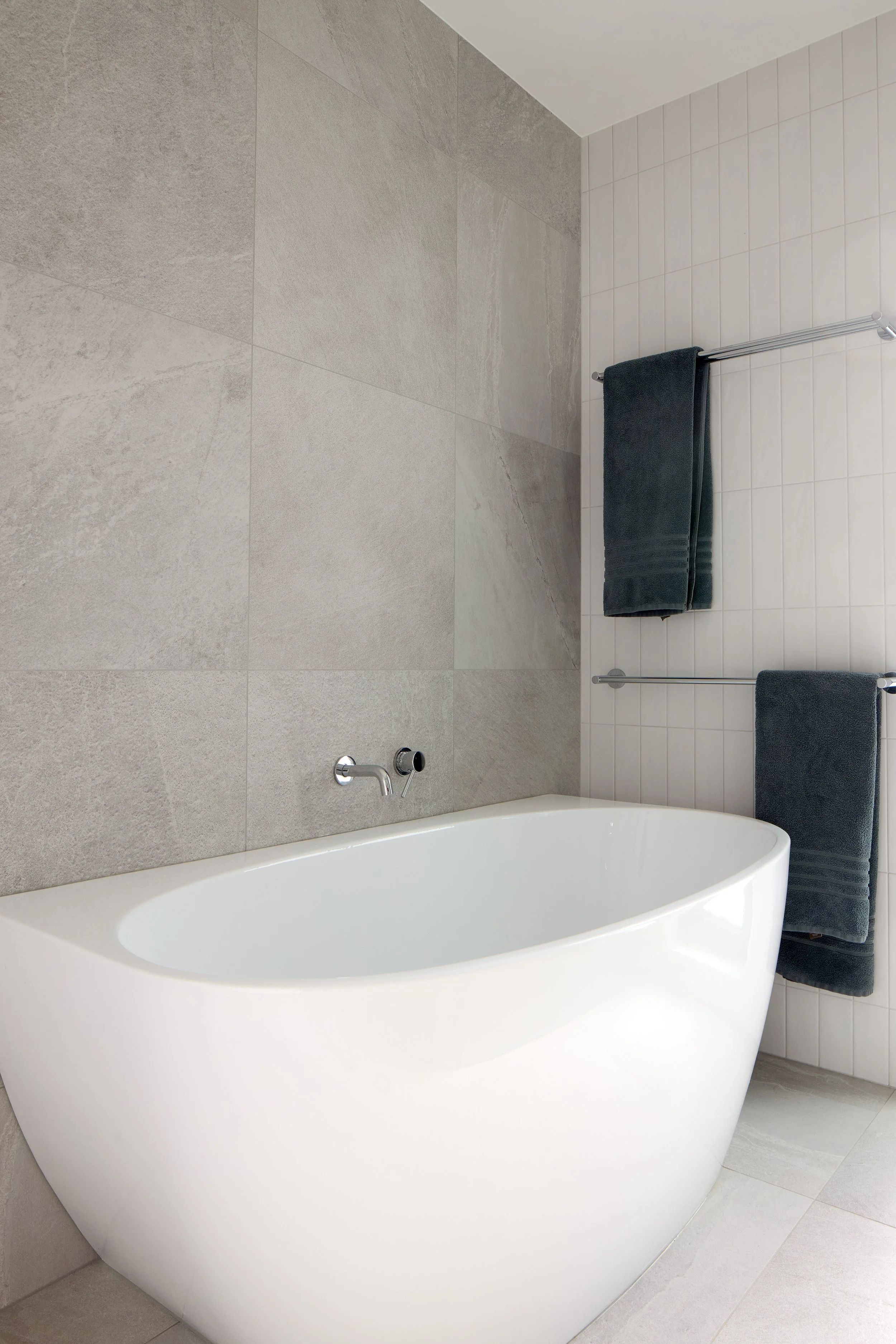 Modern bathroom with a white freestanding bathtub, gray tiled wall, and dark gray towels hanging on a silver towel rack.