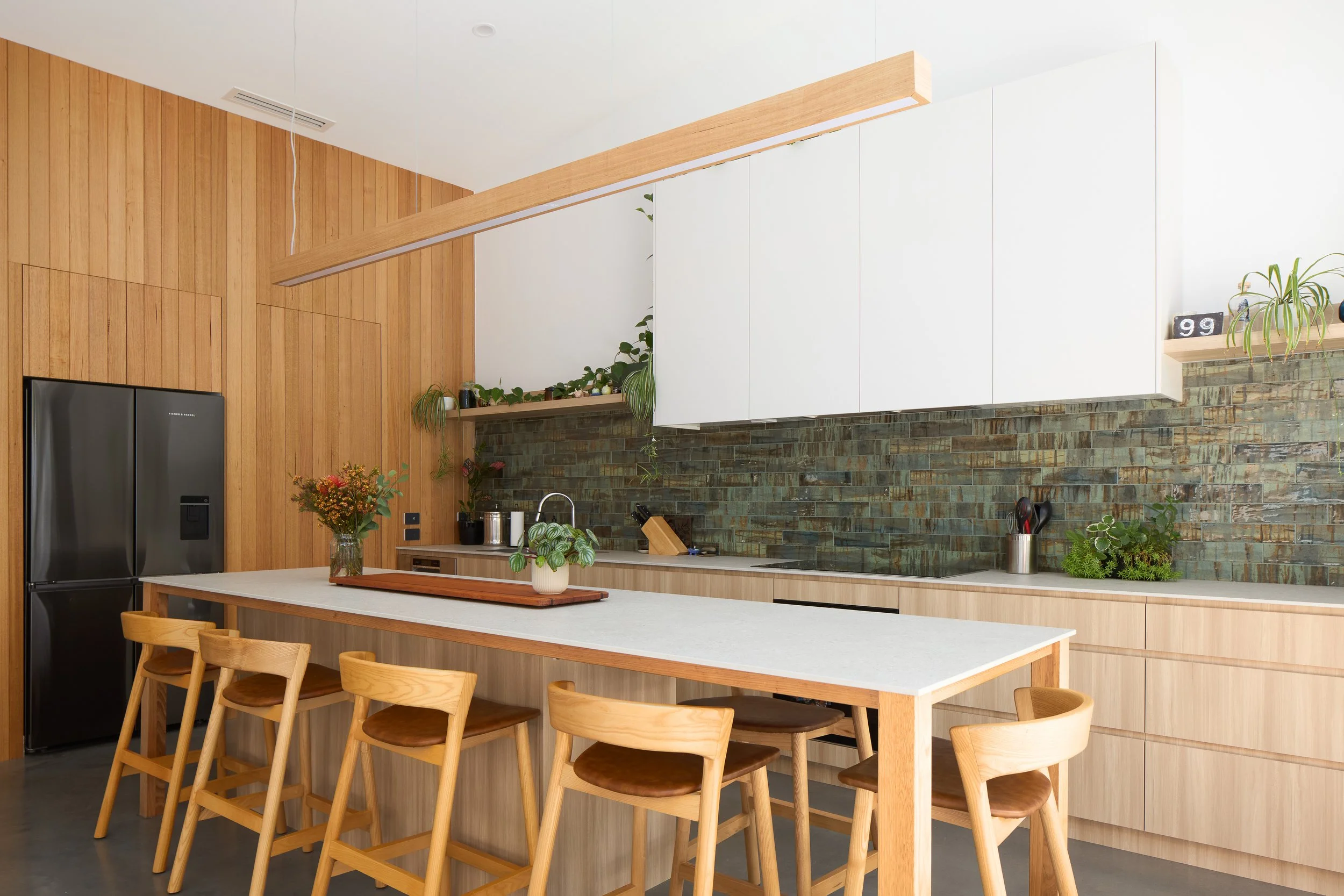 Modern kitchen with wooden walls and cabinetry, white upper cabinets, greenhouse style tile backsplash, a kitchen island with five wooden chairs, and various potted plants as decor.