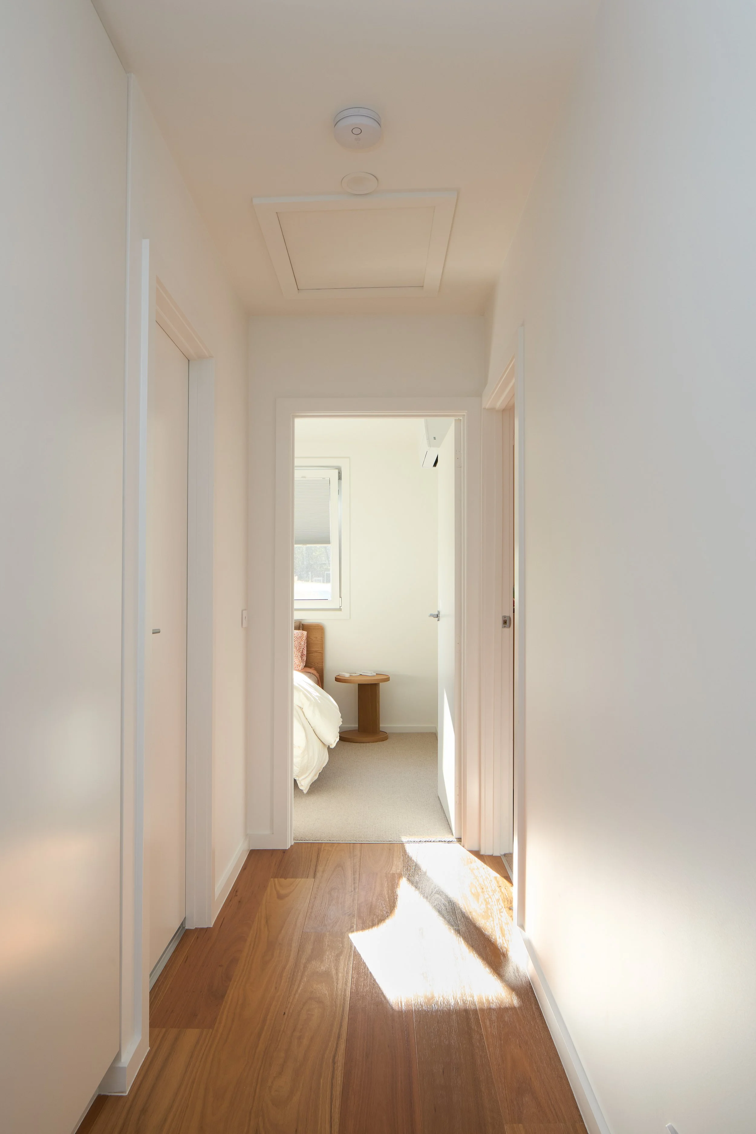 A hallway leading into a bedroom with a side table and bed near a window.