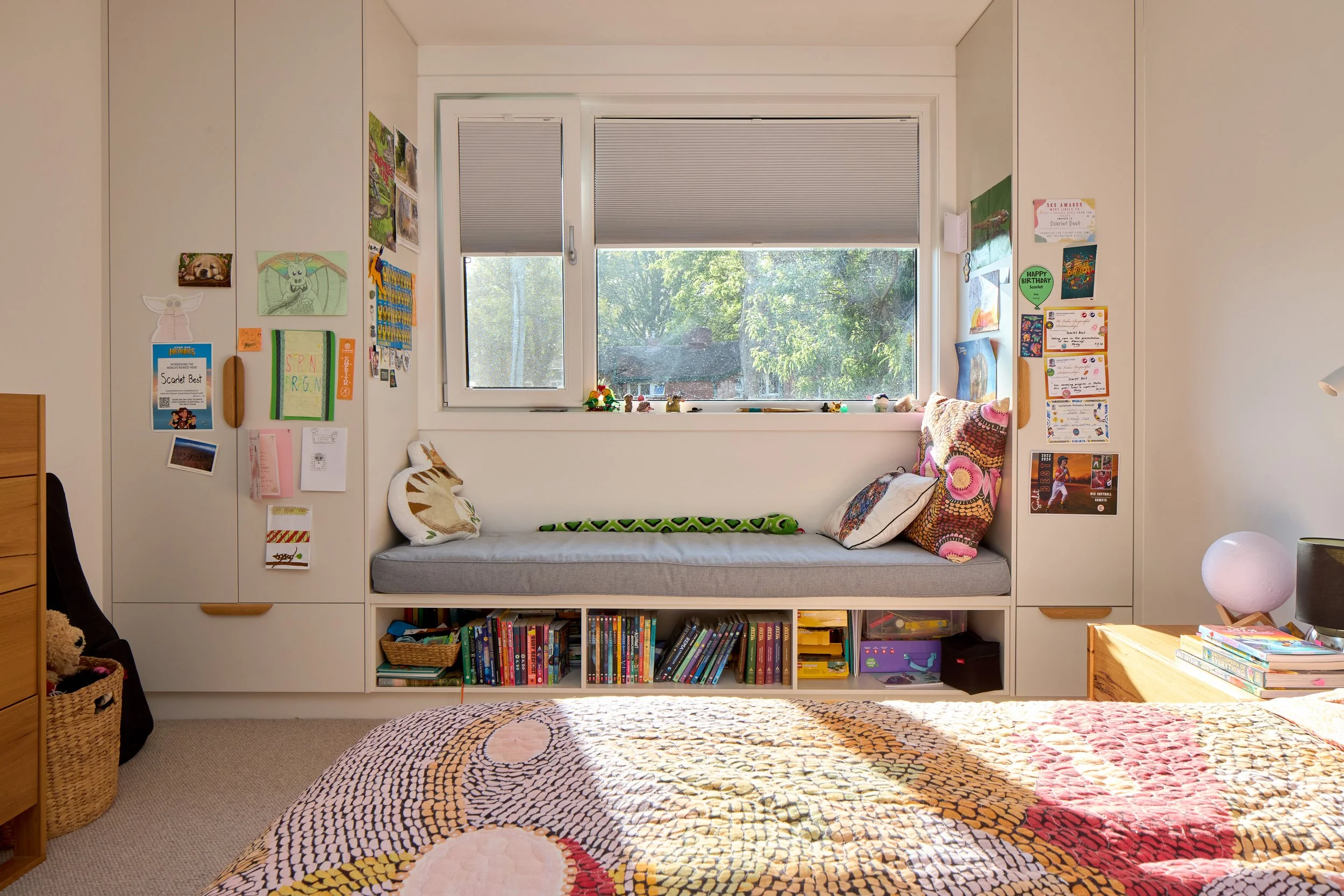 Bedroom with a large window seat, surrounded by white cabinets and walls decorated with colorful papers and pictures, a bed with a patterned quilt in the foreground, a nightstand with books and a lamp on the right side, and a basket with a teddy bear on the left.