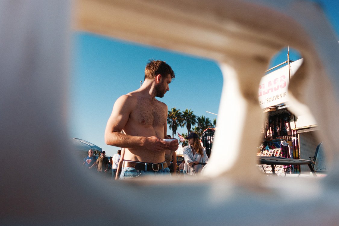 shirtless man on venice boardwalk photographed through white sidewalk sign like a frame