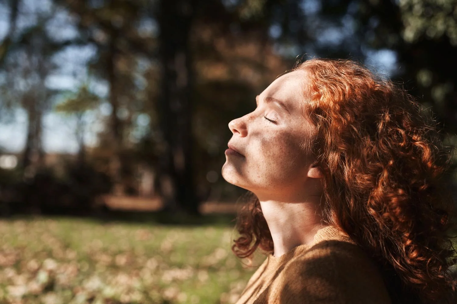 Woman with red curly hair outdoors facing the sun with eyes closed.