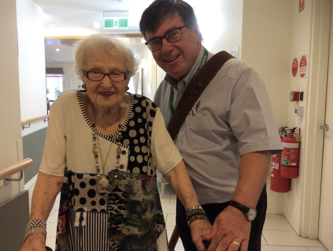 An elderly woman with white hair, glasses, and bright red lipstick standing with a middle-aged man with glasses, short dark hair, wearing a white shirt, and carrying a brown shoulder bag, inside a building.