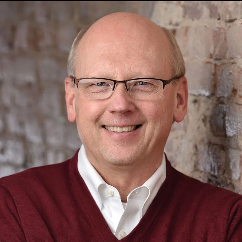 Black and white photo of a smiling man with glasses, wearing a white shirt and a dark sweater, standing against a brick wall background.