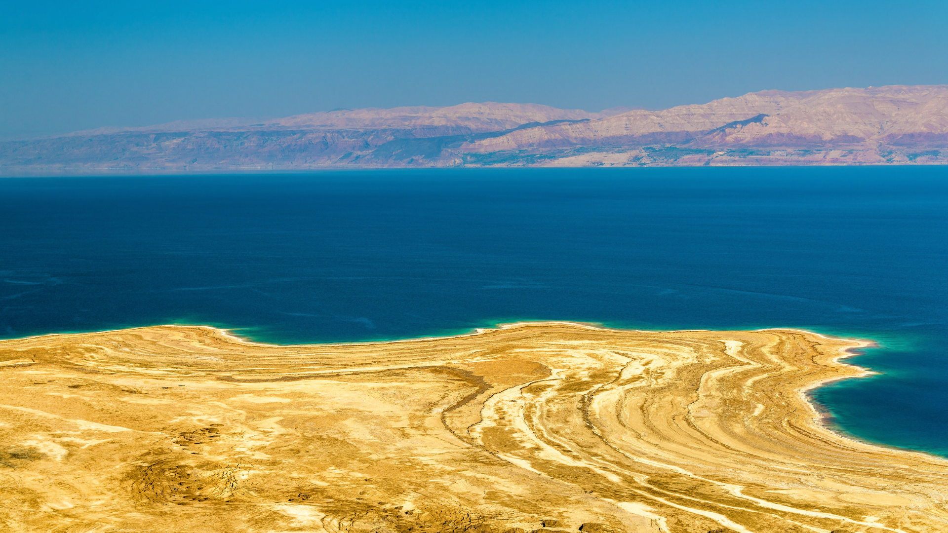 A large body of deep blue water with a landmass in the foreground featuring yellowish, textured terrain and a distant mountain range in the background under clear blue sky.