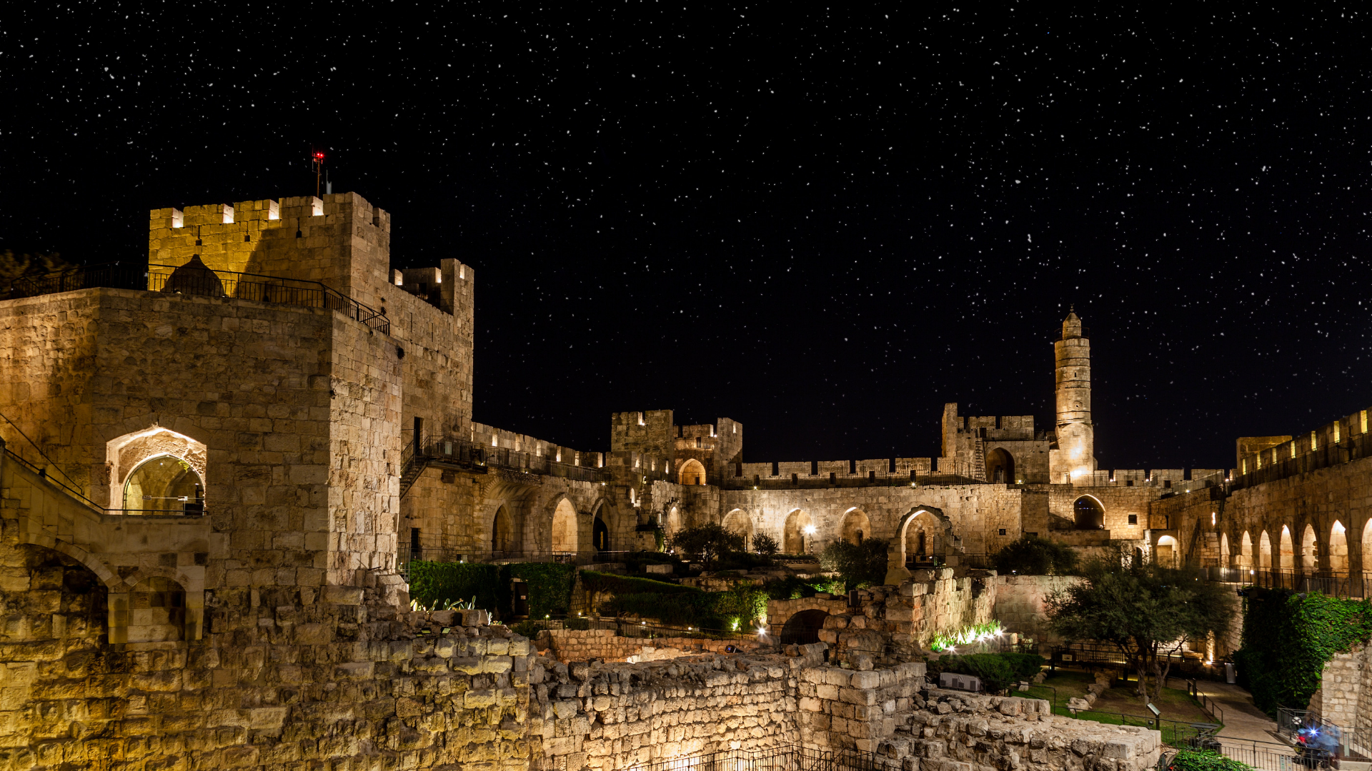 Nighttime view of an ancient stone fortress with illuminated walls and towers under a star-filled sky.