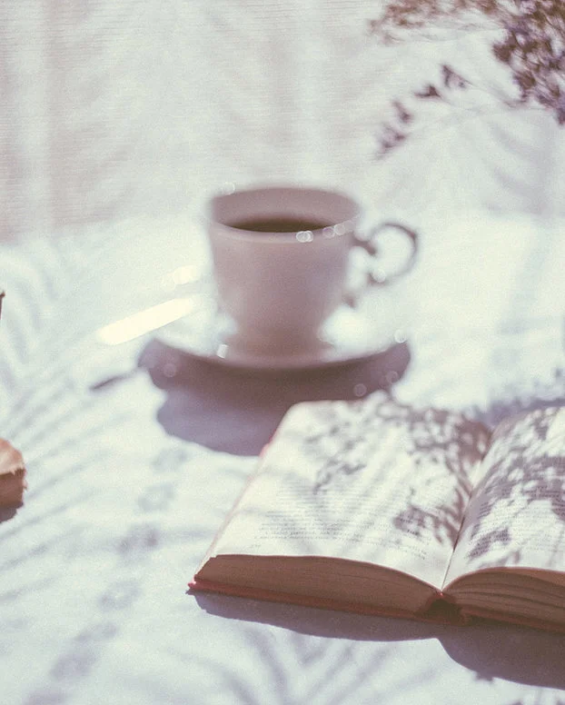 An open book, a cup of coffee on a saucer, and a spoon on a white surface with soft lighting and shadows.