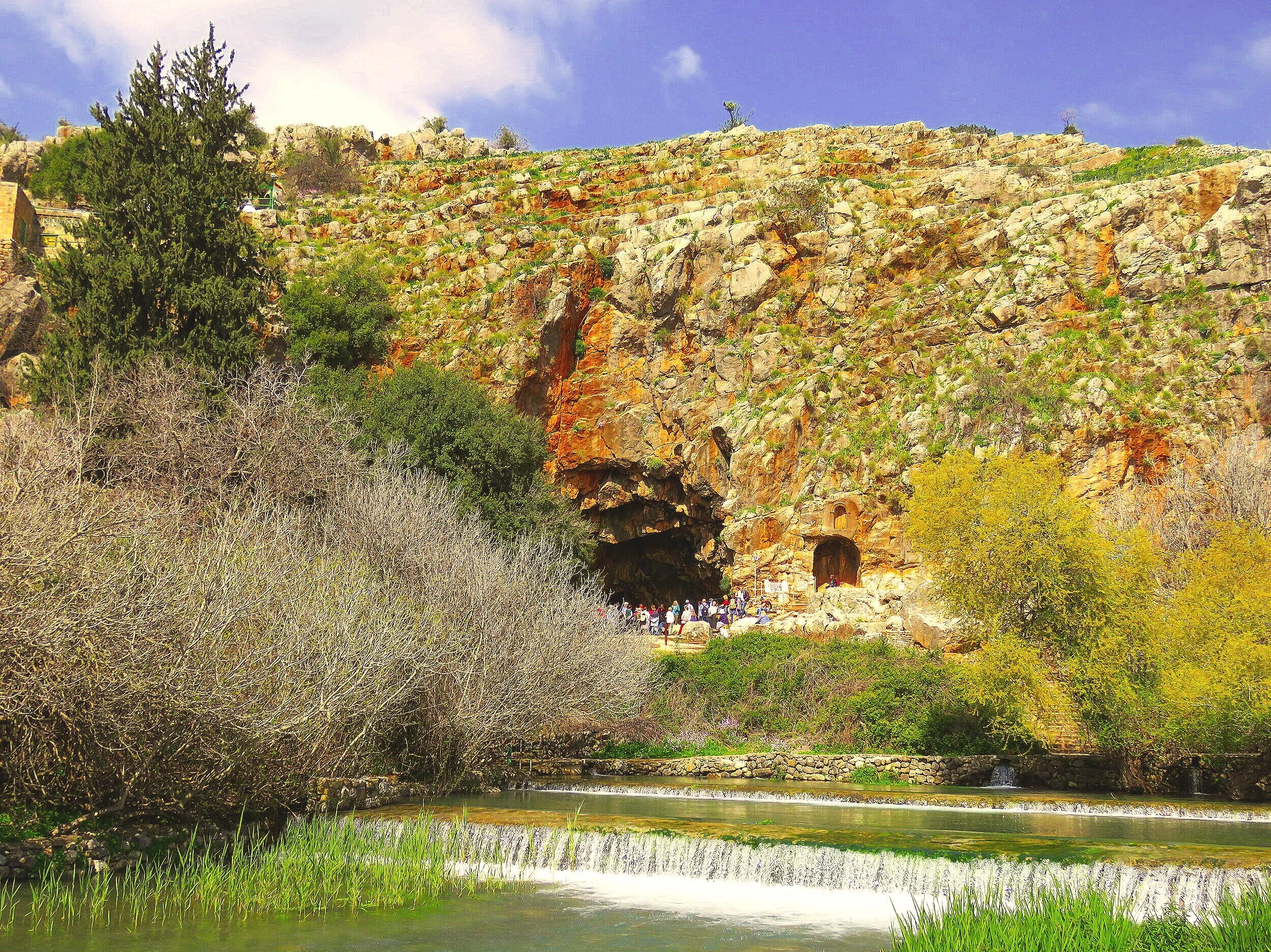 A rocky hillside with an opening leading into a cave, surrounded by trees, with a small waterfall and water below, and a group of people near the cave entrance.