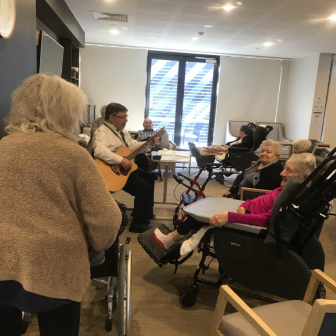Elderly residents of a nursing home watch a man playing guitar in a common room with large windows, some residents are in wheelchairs and sitting in chairs, enjoying the music.