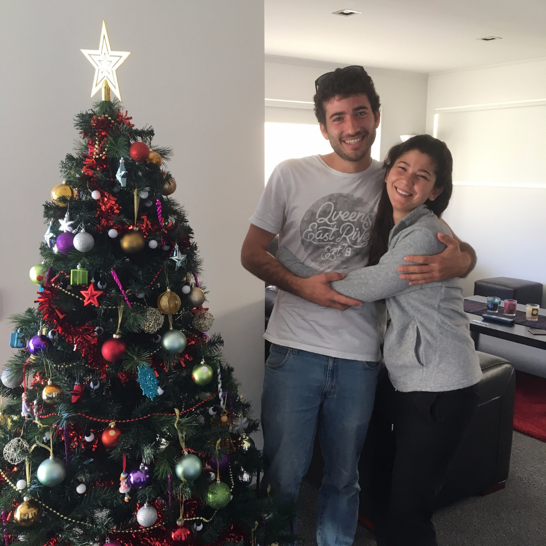 A young man and woman hugging and smiling indoors next to a decorated Christmas tree.