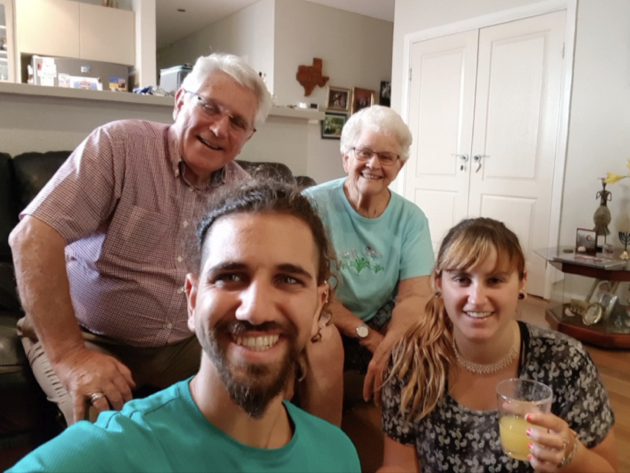 Group of five people smiling and taking a selfie in a living room. Four are sitting on a couch, including an elderly couple and two younger adults, one man and one woman. The woman is holding a yellow drink. The background shows a white cabinet, a small side table with a lamp and photo frames, and a kitchen area.