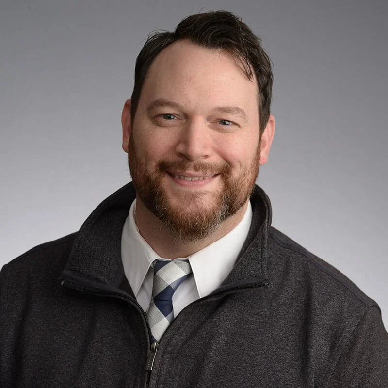Black and white portrait of a smiling man with a beard, wearing a collared shirt, tie, and a zip-up jacket.