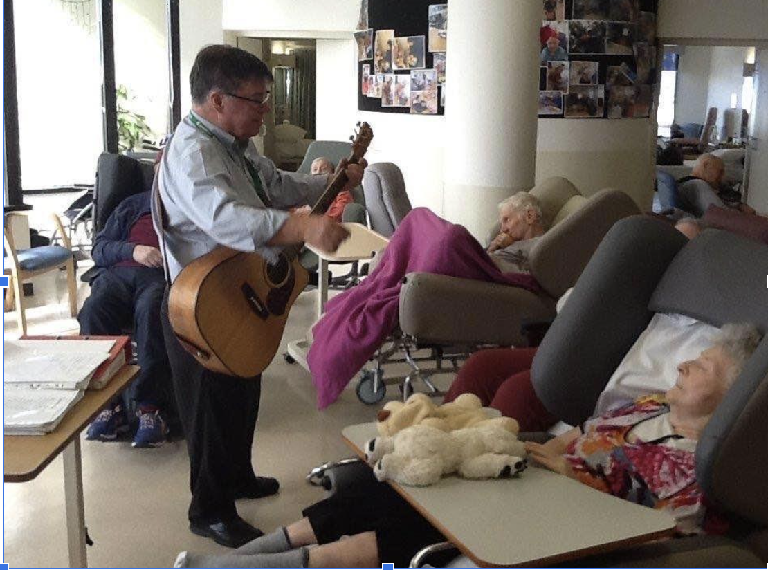 A man playing an acoustic guitar and singing to elderly residents in a care facility lounge.