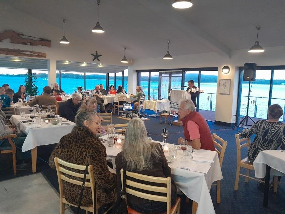People dining at tables near a large window overlooking a body of water, with a speaker and someone speaking at a podium.