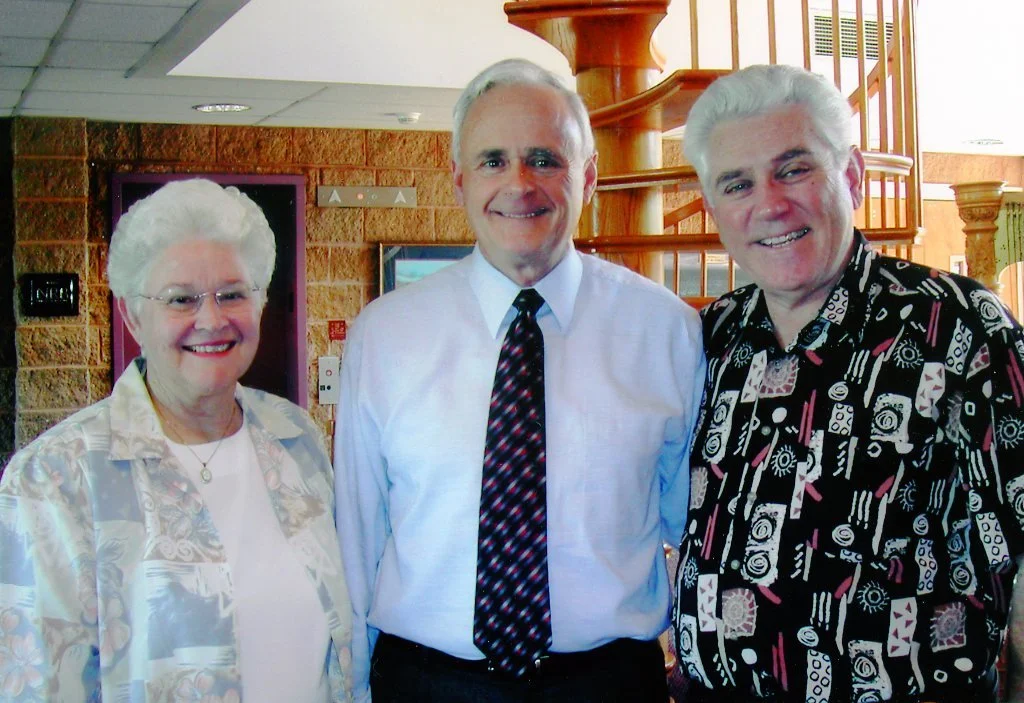 Three older adults, two men and one woman, standing together indoors in a warmly lit room, smiling at the camera.