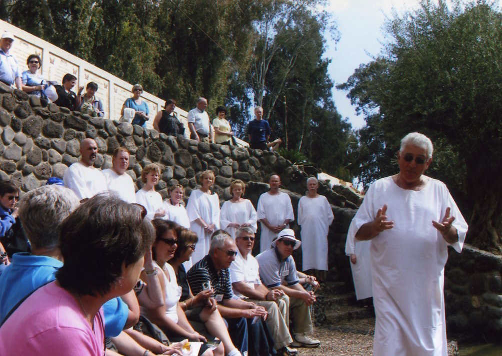 A group of people, some dressed in white, are gathered outdoors on a rocky hillside with a stone wall in the background. An elderly man in a white robe and sunglasses is speaking to the audience.