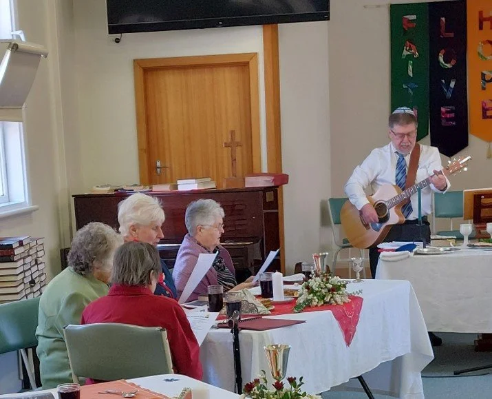 A man in a white shirt and blue tie plays a guitar and sings to four elderly women sitting at a table in a church or community room. The women are reading from paper, and the room has church banners and a cross on the wall.