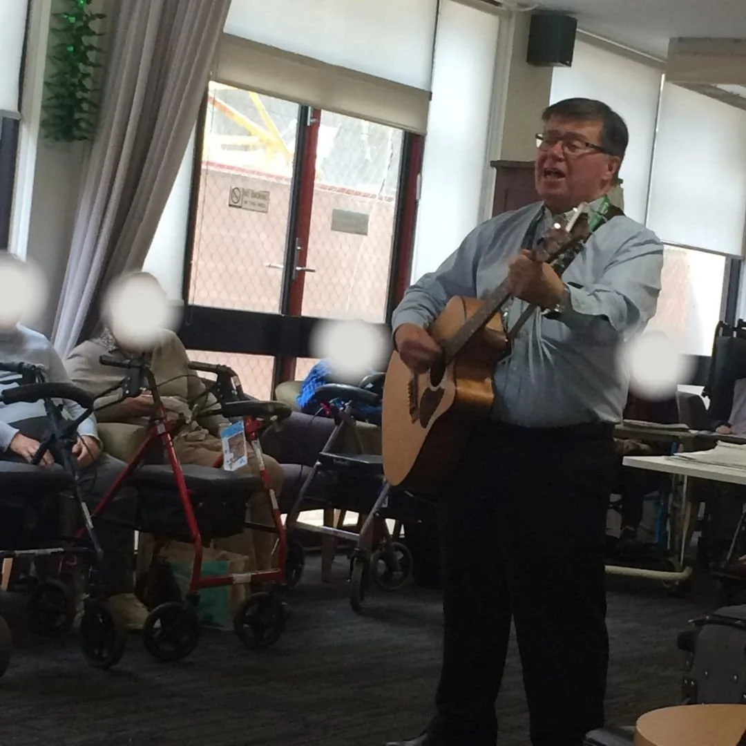 An older man with glasses singing and playing an acoustic guitar in a room with seated patients, some of whom have mobility aids, during a performance or entertainment event.