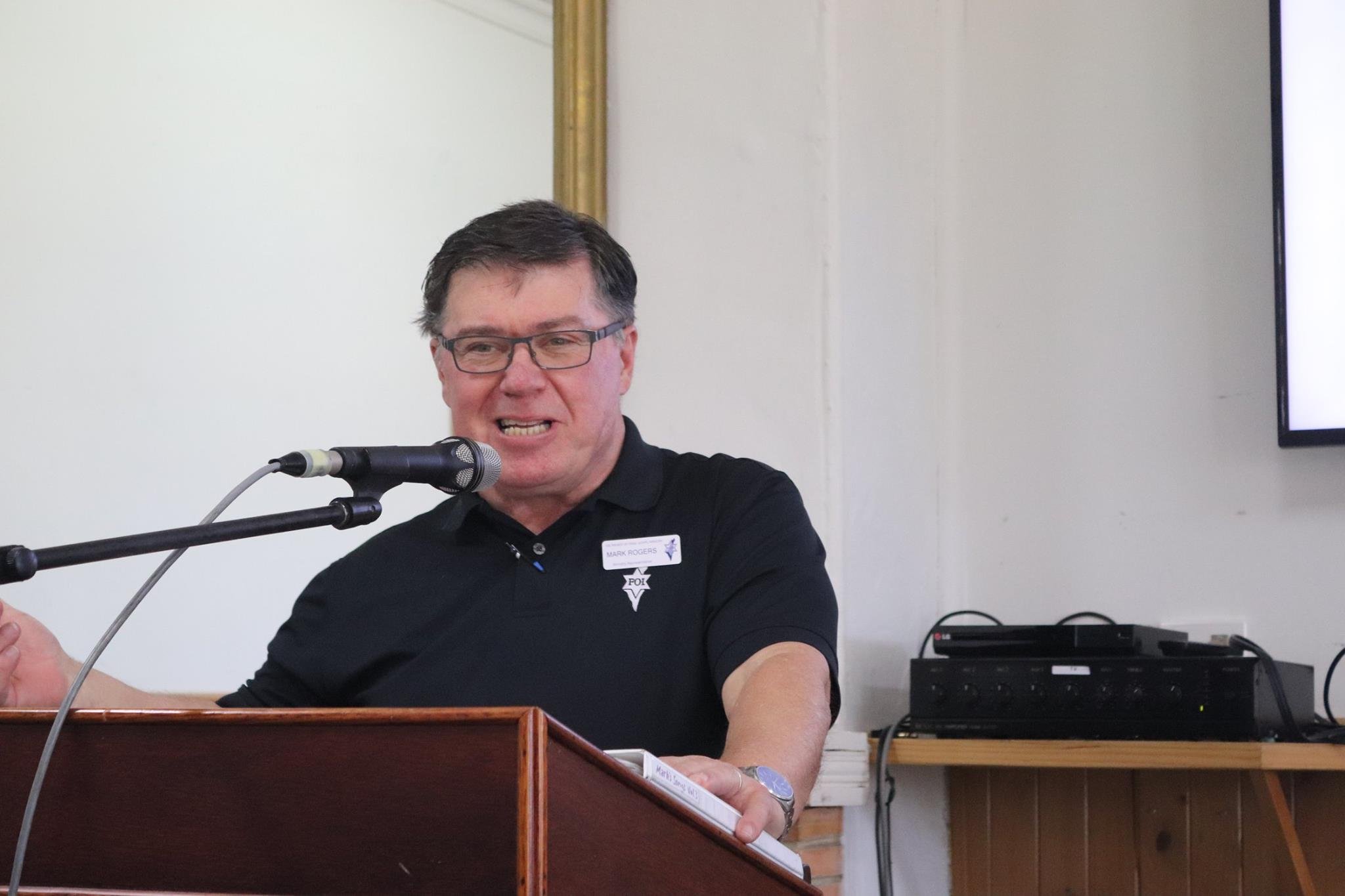 A man wearing glasses and a black shirt with a name tag speaking into a microphone at a podium, with audio equipment and a monitor in the background.